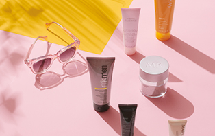 Verschillende Mary Kay producten met beschermingsfactor staan op een roze ondergrond: gezichtscrème, foundation, zonnecrème. Daarnaast ligt een roze zonnebril.