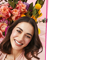 Een vrouw met bruin haar en een stralende teint glimlacht naar de camera. Op de achtergrond is een boeket bloemen te zien.
