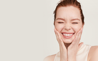 Eine Frau mit hellem Teint fasst sich mit beiden Händen ans Gesicht, hat die Augen geschlossen und lächelt. Um sie herum sieht man gezeichnete Inhaltsstoffe von Hautpflegeprodukten.