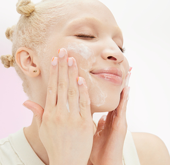 Een jonge vrouw heeft haar ogen gesloten en brengt met beide handen een scrub aan op haar gezicht