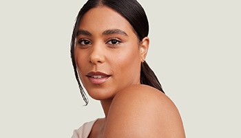 Een lachende vrouw met bruin haar, een stralende huid en gesloten ogen grijpt met één hand naar haar gezicht