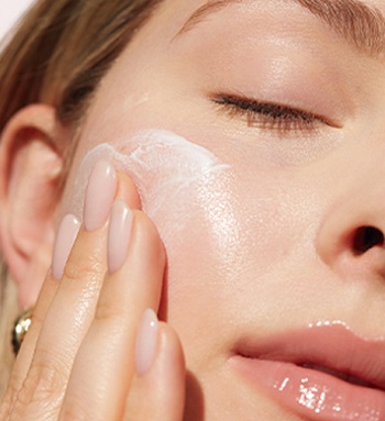 Close-up van het gezicht van een jonge vrouw met gesloten ogen, die met één hand TimeWise Antioxidant Moisturizer uit de TimeWise "Wonder-set" op haar gezicht aanbrengt.