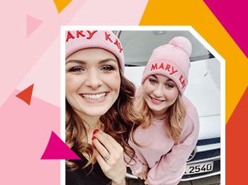 Twee stralende vrouwen met pinke Mary Kay mutsen staan voor een witte auto.