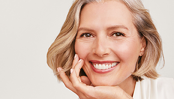 Een blonde vrouw met een rijpe huid houdt haar kin in haar hand en glimlacht naar de camera