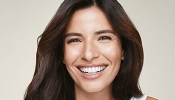 Een vrouw van in de 30 met donker, lang haar en een stralende, heldere huid lacht frontaal in de camera