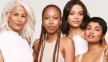 Vier vrouwen van verschillende leeftijden met verschillende huidskleuren, huidskleuren en haarkleuren kijken recht in de camera. Ze hebben allemaal een stralende, heldere huid.