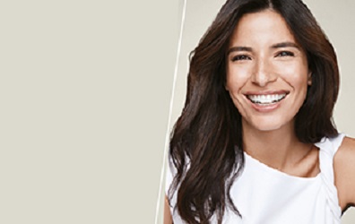 Een vrouw van in de 30 met donker, lang haar en een stralende, heldere huid lacht frontaal in de camera