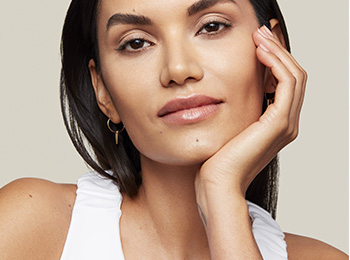 Een jonge vrouw met donker haar en een stralend mooie huid kijkt in de camera en steunt haar gezicht met haar hand