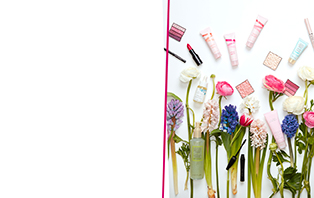 Mary Kay cadeau-ideeën voor Pasen 2022: Mary Kay producten die geweldig zijn om cadeau te geven zweven boven een kleurrijke lenteweide met bloeiende bloemen - lippenstift, blush, parfum, handcrème, oogschaduw, gezichtsolie, highlighter, en meer