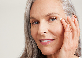 Een vrouw van boven de 60 met een rijpe, verzorgde huid en een kort kapsel lacht frontaal in de camera en raakt met haar hand haar huid aan