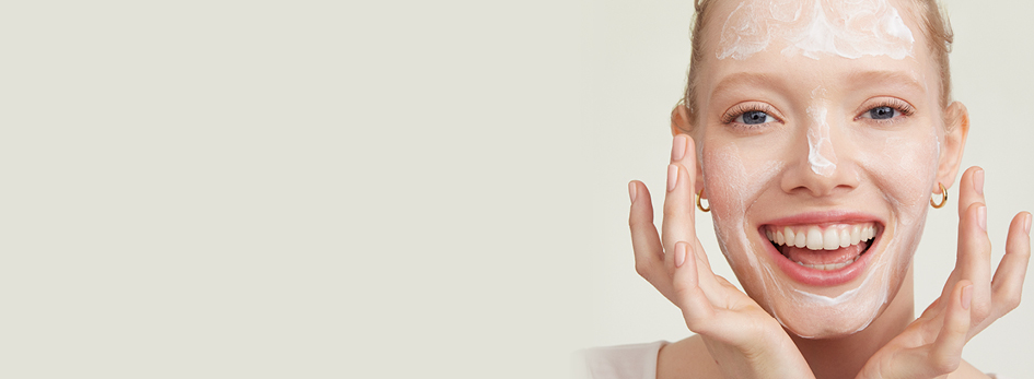 Een vrouw met een stralende glimlach brengt met beide handen een scrub aan op haar gezicht