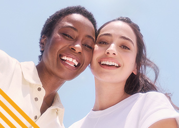 Een vrouw met een donkere huidskleur en een vrouw met een medium huidskleur staan voor een lichtblauwe achtergrond. De vrouwen glimlachen allebei en hebben een positieve uitstraling.