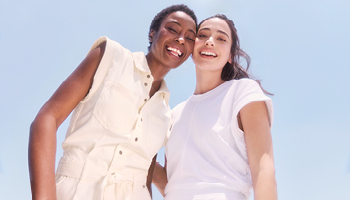 Een vrouw met een donkere huidskleur en een vrouw met een medium huidskleur staan voor een lichtblauwe achtergrond. De vrouwen glimlachen allebei en hebben een positieve uitstraling.
