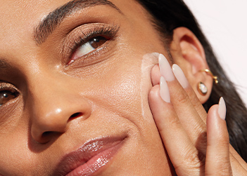 Close-up van een vrouw met donkere huid die zelfverzekerd opzij kijkt terwijl ze gezichtsverzorging op haar wang aanbrengt