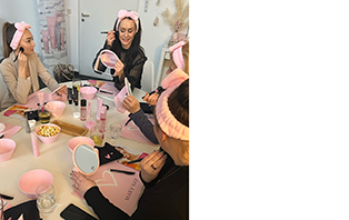 Verschillende vrouwen met roze hoofdbanden in hun haar zitten aan een tafel tijdens een Mary Kay party met een zelfstandige schoonheidsconsulente, waarop enkele Mary Kay producten, spiegels etc. te zien zijn.