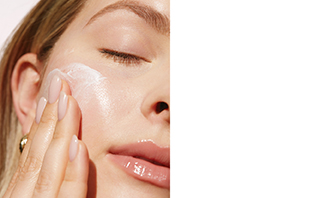 Close-up van het gezicht van een vrouw die haar ogen heeft gesloten en met één hand huidverzorgingscrème in haar wangen masseert.